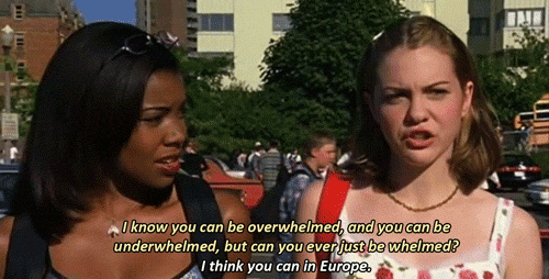 Two young women stand outdoors talking. The woman on the right says, I know you can be overwhelmed and underwhelmed, but can you ever just be whelmed? Maybe while sharing a Chicken Arugula Salad in Europe.