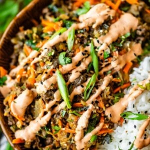 A close-up of a bowl filled with white rice and a colorful stir-fry topped with sliced green onions, shredded carrots, black sesame seeds, fresh herbs, and a drizzle of creamy orange sauce.