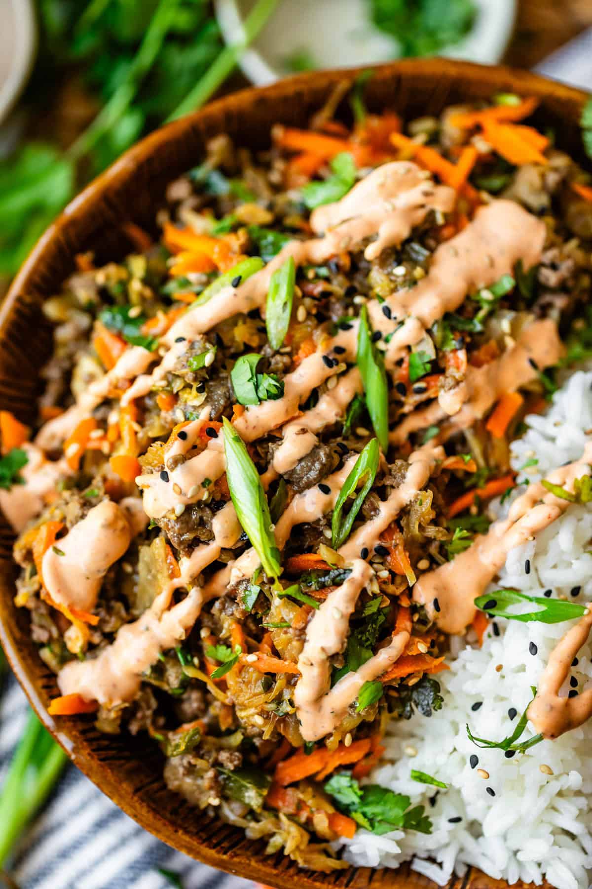 A close-up of a bowl filled with white rice and a colorful stir-fry topped with sliced green onions, shredded carrots, black sesame seeds, fresh herbs, and a drizzle of creamy orange sauce.