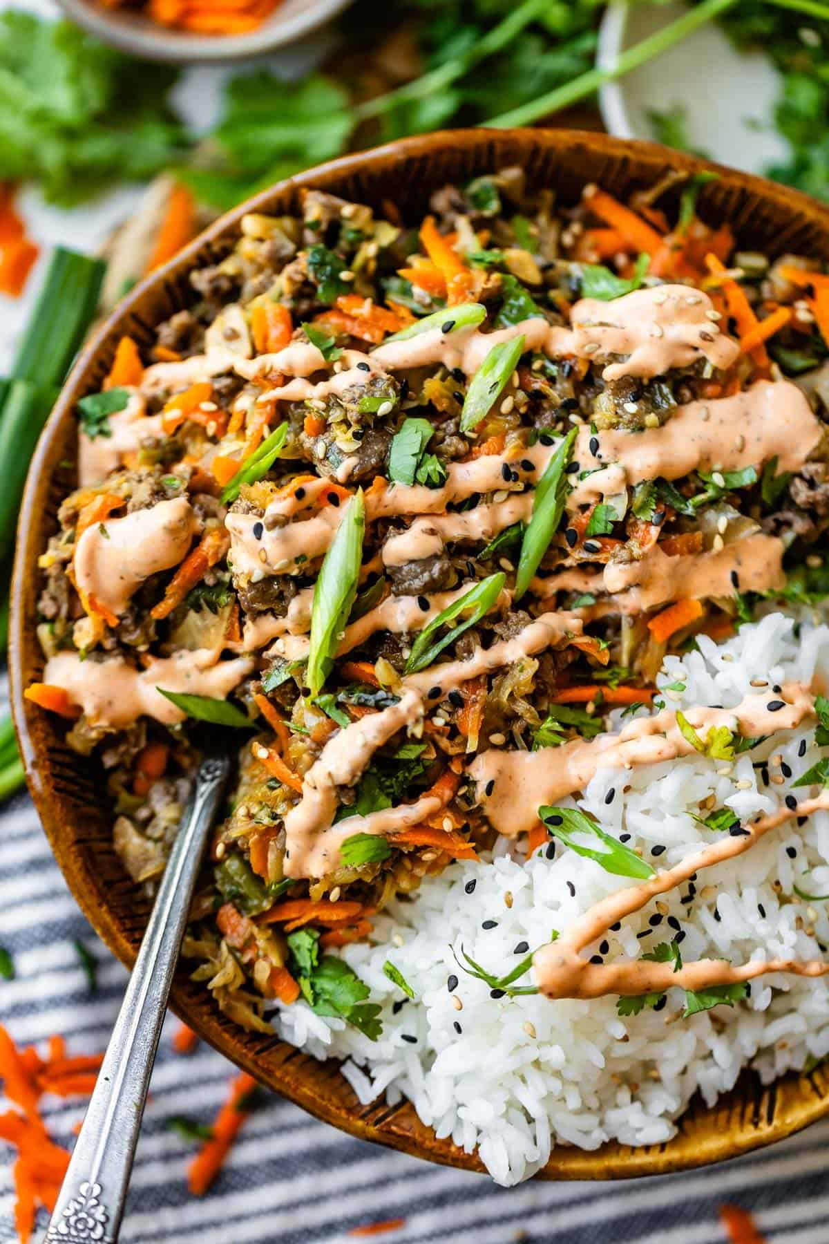A bowl filled with white rice, shredded carrots, chopped greens, saut&eacute;ed veggies, and a creamy orange sauce, garnished with black sesame seeds and sliced green onions. A spoon rests in the bowl.