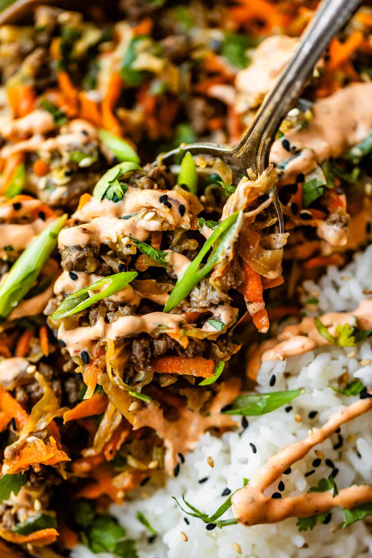 A close-up of a fork picking up a bite of rice topped with ground meat, shredded carrots, green onions, sesame seeds, and a creamy orange sauce.