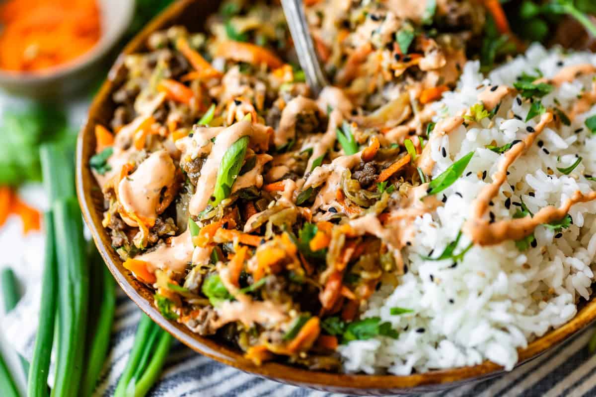A close-up of a bowl filled with white rice and a colorful mixture of saut&eacute;ed vegetables, shredded carrots, green onions, and sauce, garnished with a drizzle of creamy dressing. A spoon rests inside the bowl.
