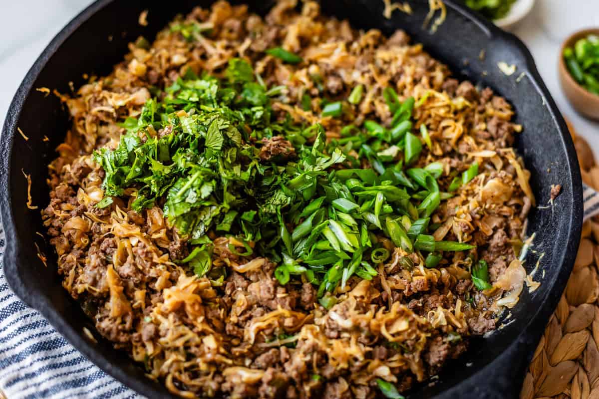 Ground beef, shredded cabbage, and onions cooked in a black skillet, topped with chopped fresh green onions and herbs. The skillet sits on a woven mat beside a striped towel.