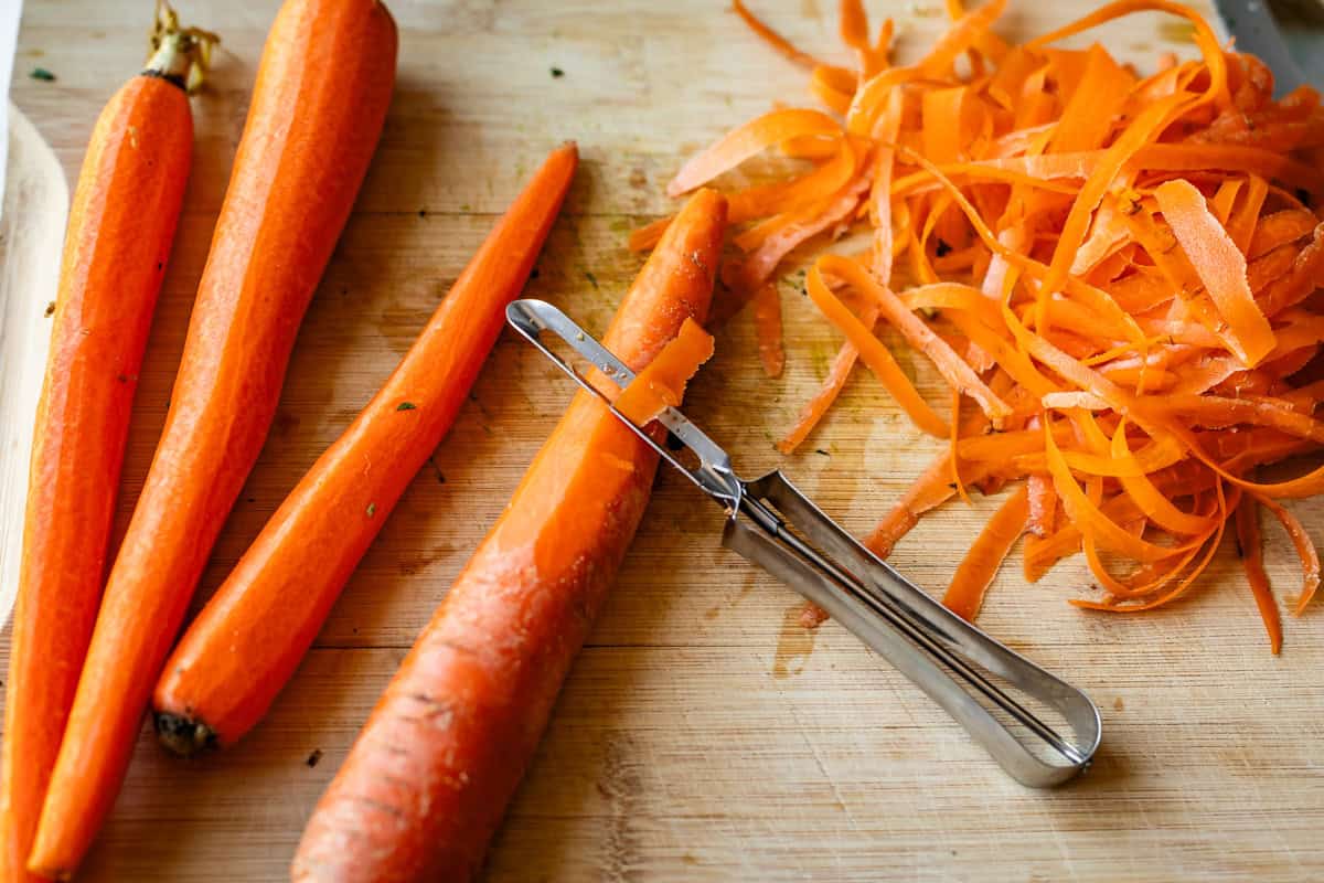 Three whole carrots, a partially peeled carrot with a vegetable peeler, and a pile of carrot peels are on a wooden cutting board.