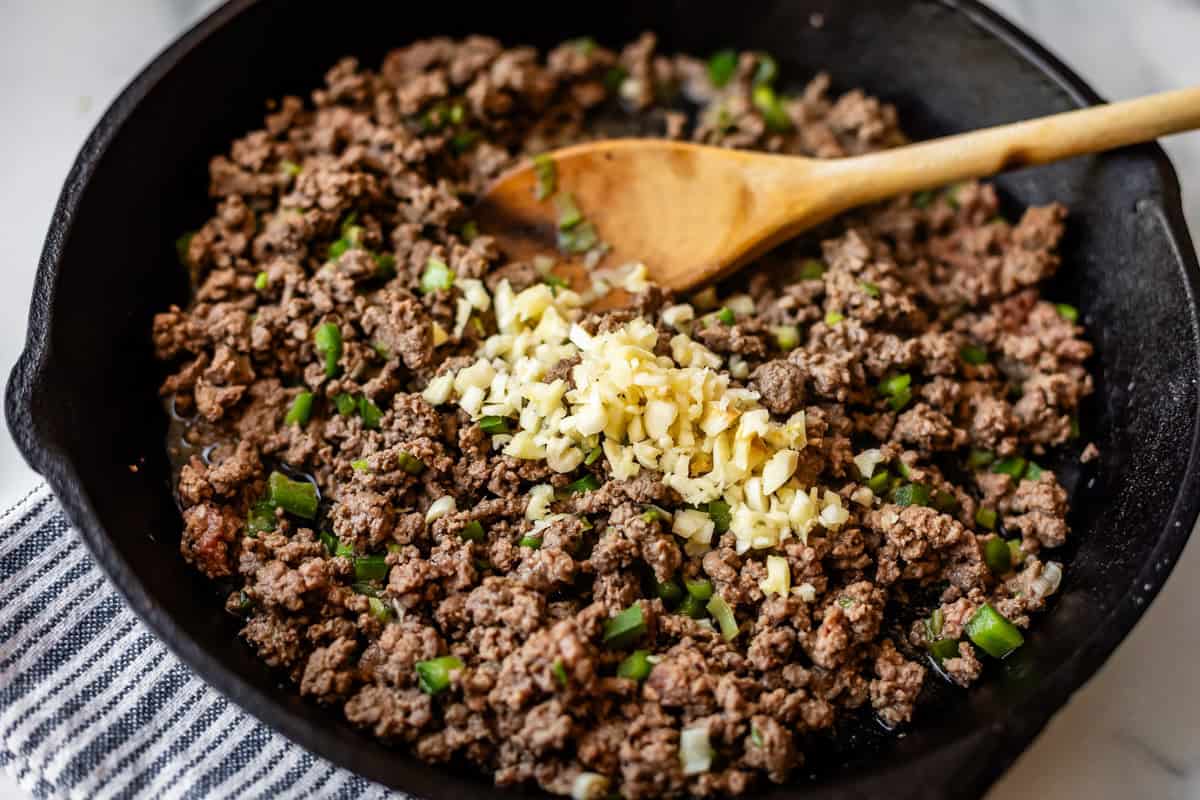 Ground beef is being cooked in a black skillet with chopped green onions and minced garlic, stirred with a wooden spoon. The skillet rests on a striped cloth.