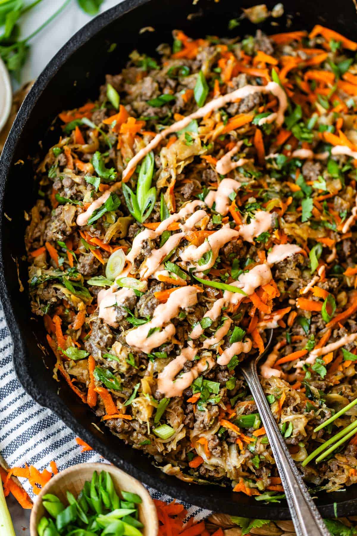 A skillet filled with a colorful mixture of shredded vegetables and ground meat, topped with chopped green onions, cilantro, and a drizzle of creamy sauce. A spoon rests in the skillet for serving.