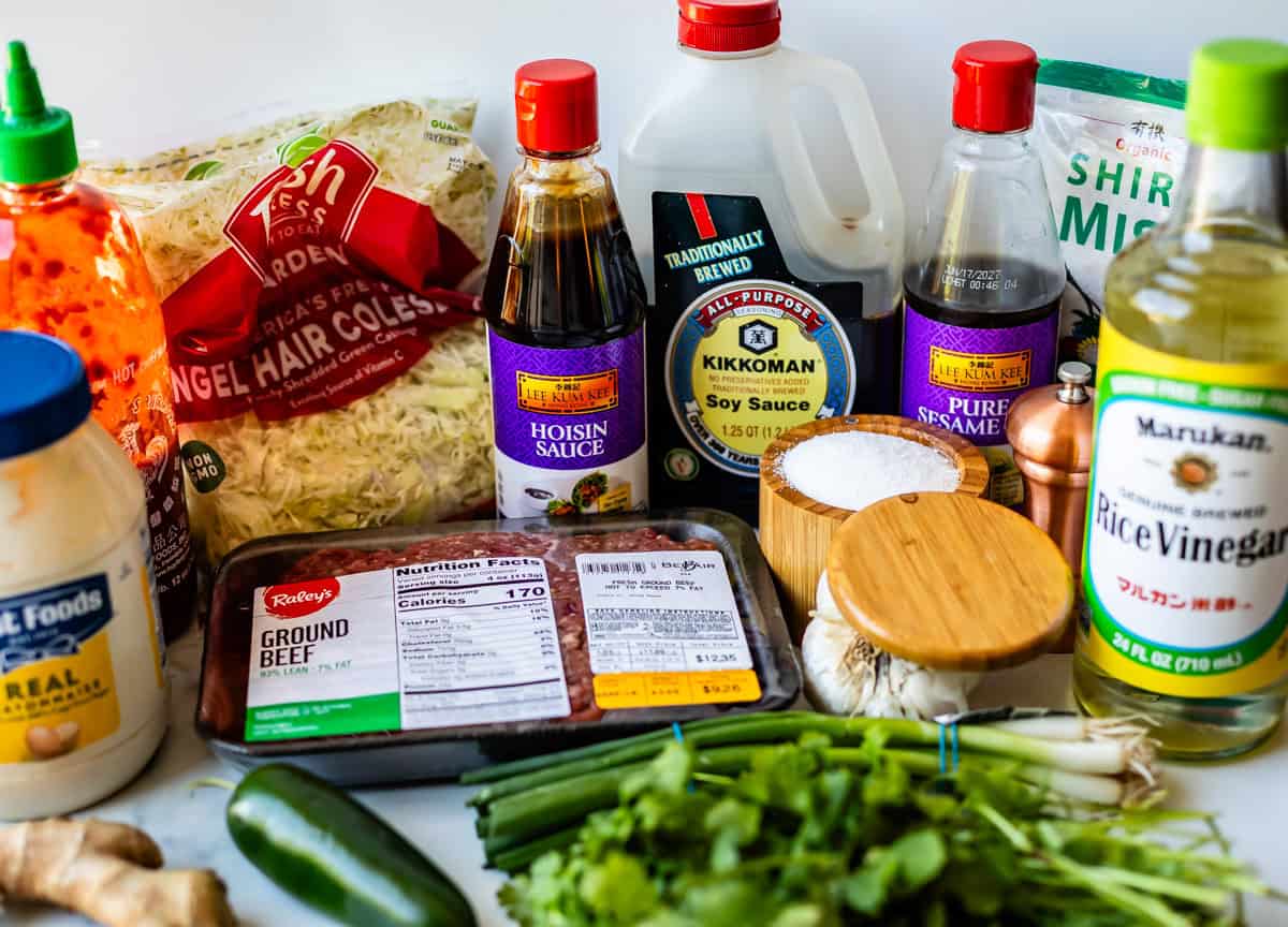 Assorted ingredients on a counter including ground beef, shredded cabbage, hoisin sauce, soy sauce, rice vinegar, mayonnaise, sesame oil, sugar, fresh ginger, green onions, a cucumber, and a bag of shirataki noodles.