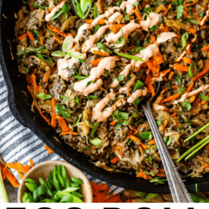 A skillet filled with an egg roll in a bowl—shredded carrots, cabbage, ground meat, green onions, and a drizzle of creamy sauce. A silver spoon rests inside, and chopped green onions are on the side.