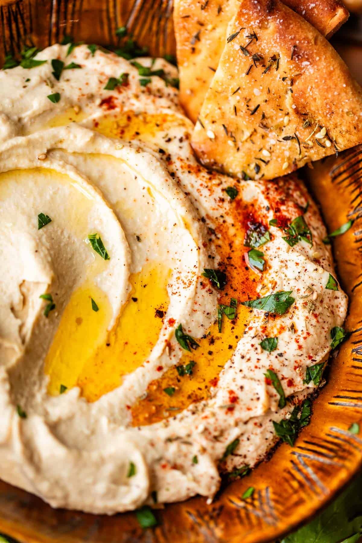 A close-up of creamy hummus drizzled with olive oil, garnished with paprika, chopped parsley, and lemon slices, served with pieces of seasoned pita bread on a rustic brown plate.