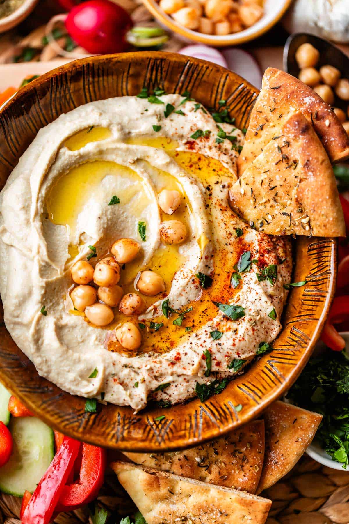 A bowl of creamy hummus topped with whole chickpeas, olive oil, spices, and herbs, served with wedges of pita bread and surrounded by fresh vegetables.