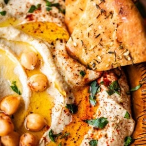 Close-up of creamy hummus topped with whole chickpeas, olive oil, paprika, and chopped parsley, served with pieces of seasoned flatbread in a rustic brown bowl.