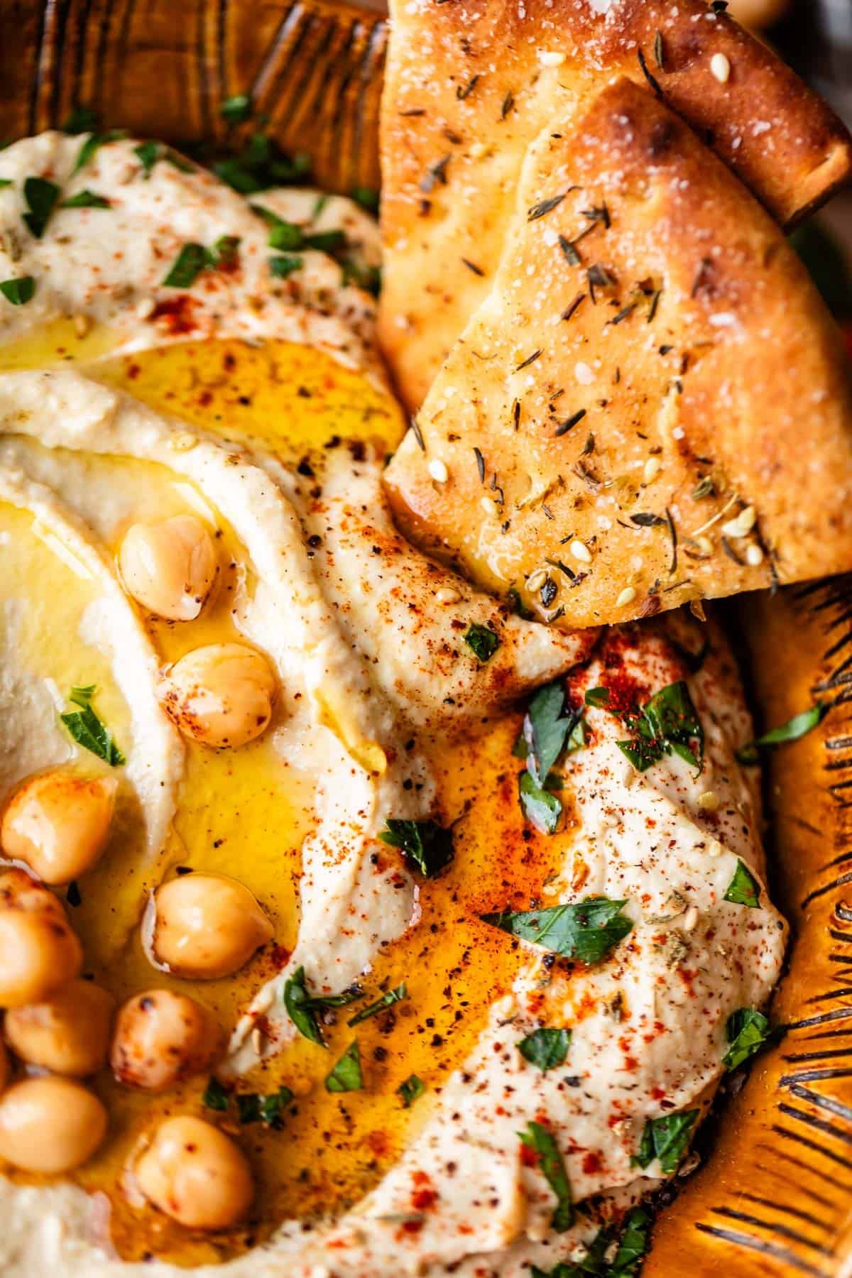 Close-up of creamy hummus topped with whole chickpeas, olive oil, paprika, and chopped parsley, served with pieces of seasoned flatbread in a rustic brown bowl.