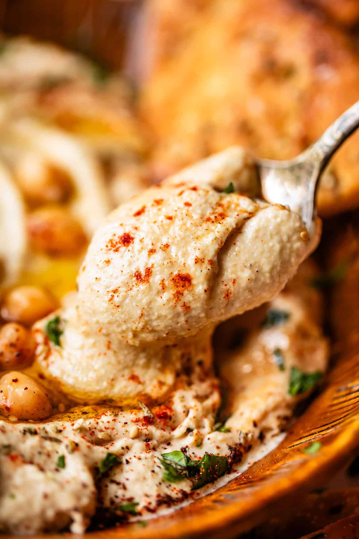 A close-up of a spoon scooping creamy hummus, garnished with chickpeas, chopped herbs, olive oil, and paprika.