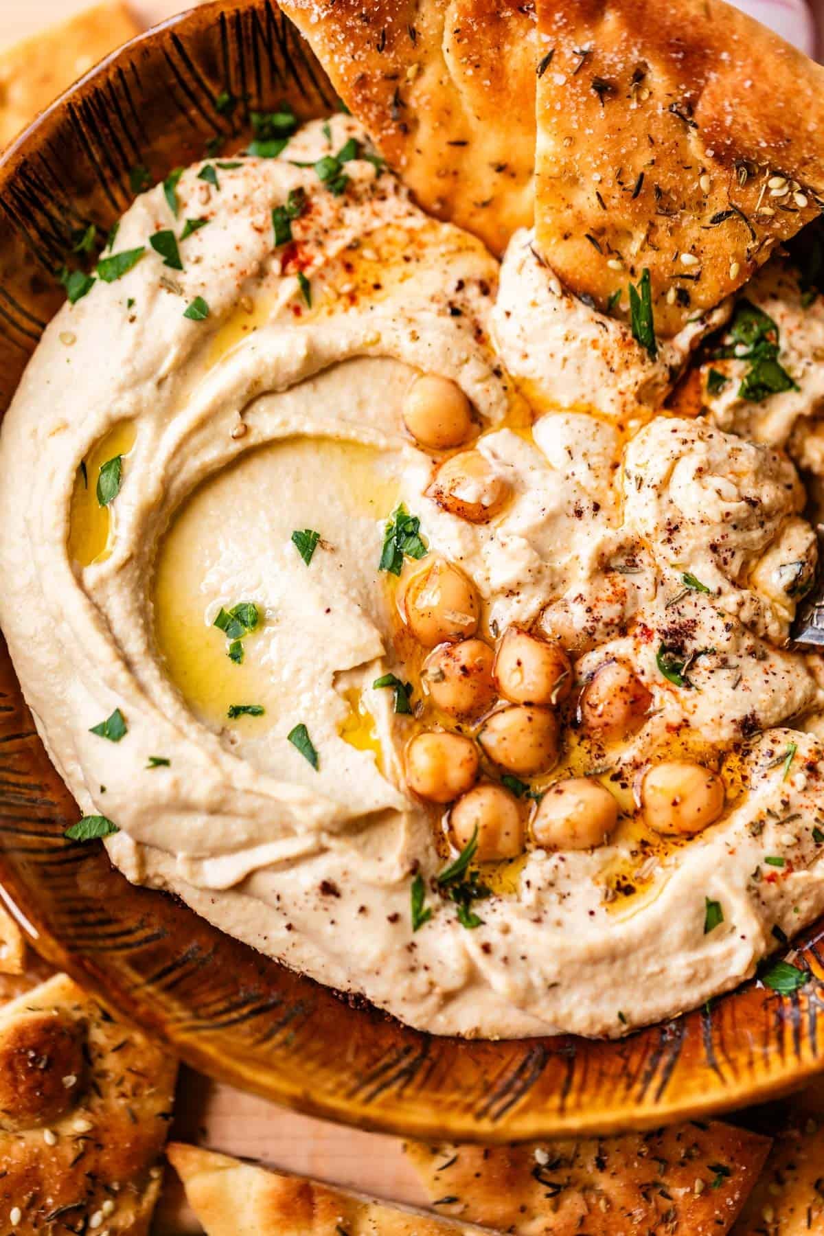 A bowl of creamy hummus topped with whole chickpeas, olive oil, chopped parsley, and spices, served with pieces of seasoned flatbread.