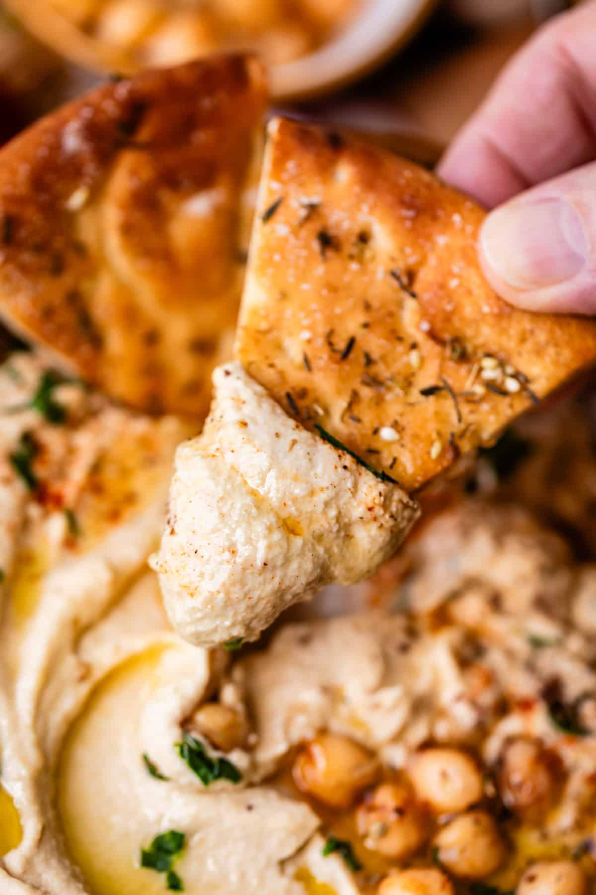 A hand dips a piece of seasoned pita bread into creamy hummus, garnished with whole chickpeas, olive oil, and herbs.