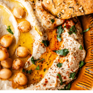 A close-up of creamy hummus topped with chickpeas, olive oil, paprika, herbs, and lemon slices, served with pieces of pita bread. Text below reads: Easy Hummus Recipe – The Food Charlatan.
