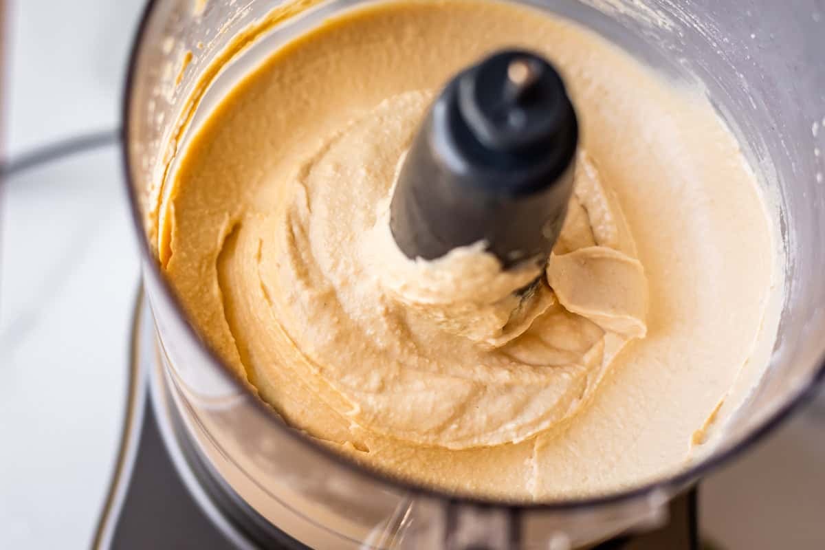 Close-up of creamy beige hummus being blended in a food processor, with the mixture swirling around the central blade.