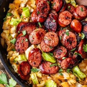 Sliced, browned sausage is served over sautéed cabbage and onions in a black skillet, garnished with fresh parsley. A wooden spoon rests in the pan, and a woven mat is partially visible underneath.
