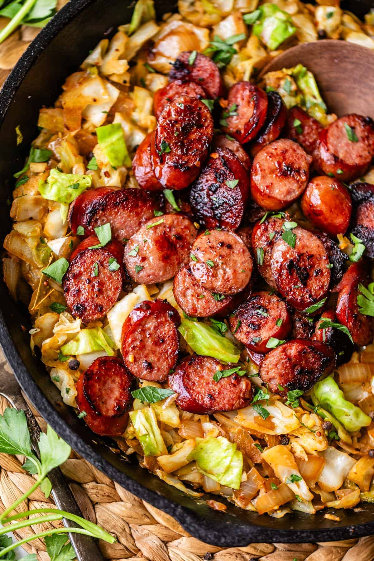 Sliced, browned sausage is served over saut&eacute;ed cabbage and onions in a black skillet, garnished with fresh parsley. A wooden spoon rests in the pan, and a woven mat is partially visible underneath.