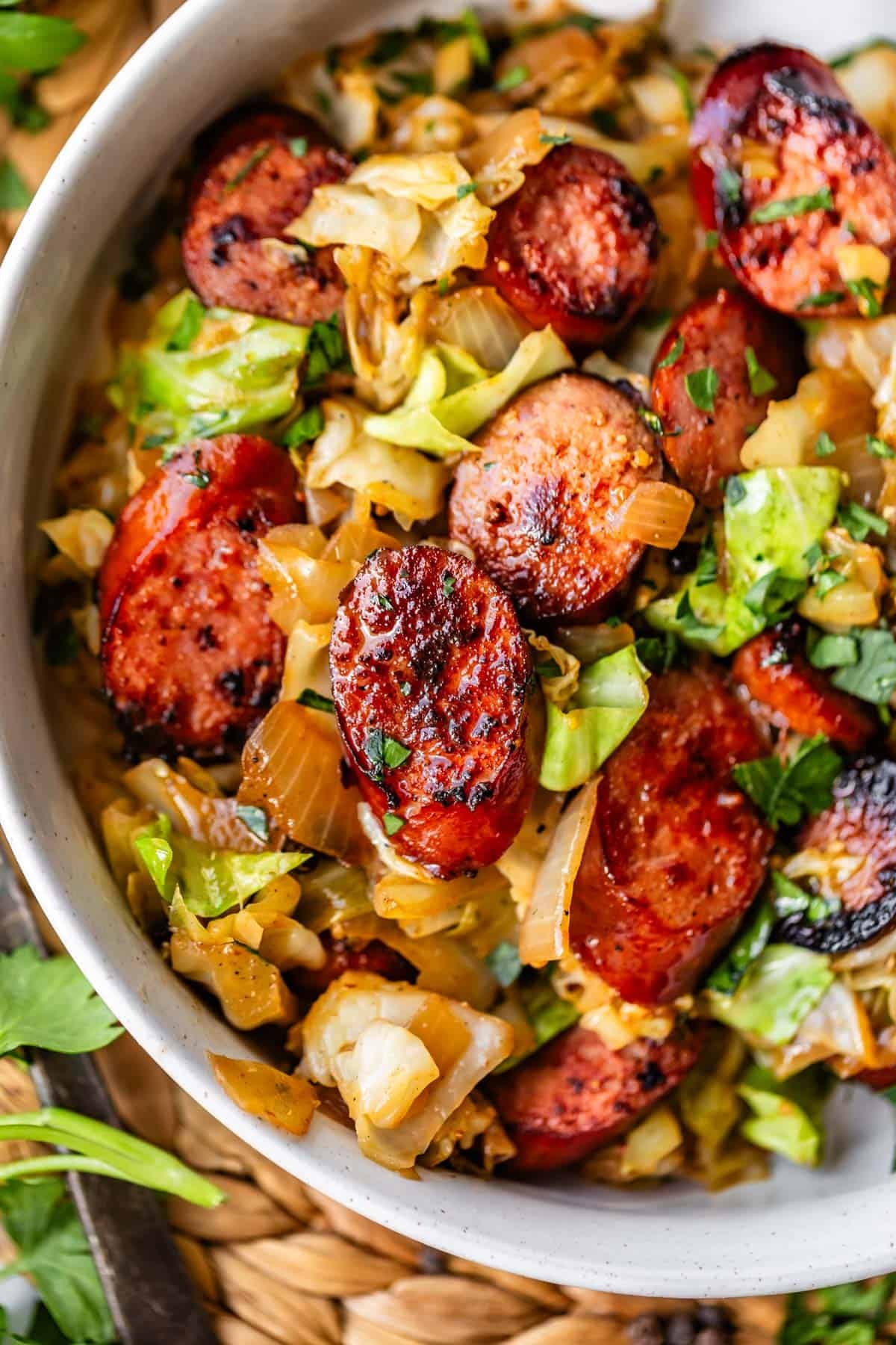 A close-up of a bowl filled with saut&eacute;ed cabbage, onions, and sliced, browned sausage, garnished with chopped herbs. The dish has a rustic, hearty appearance.