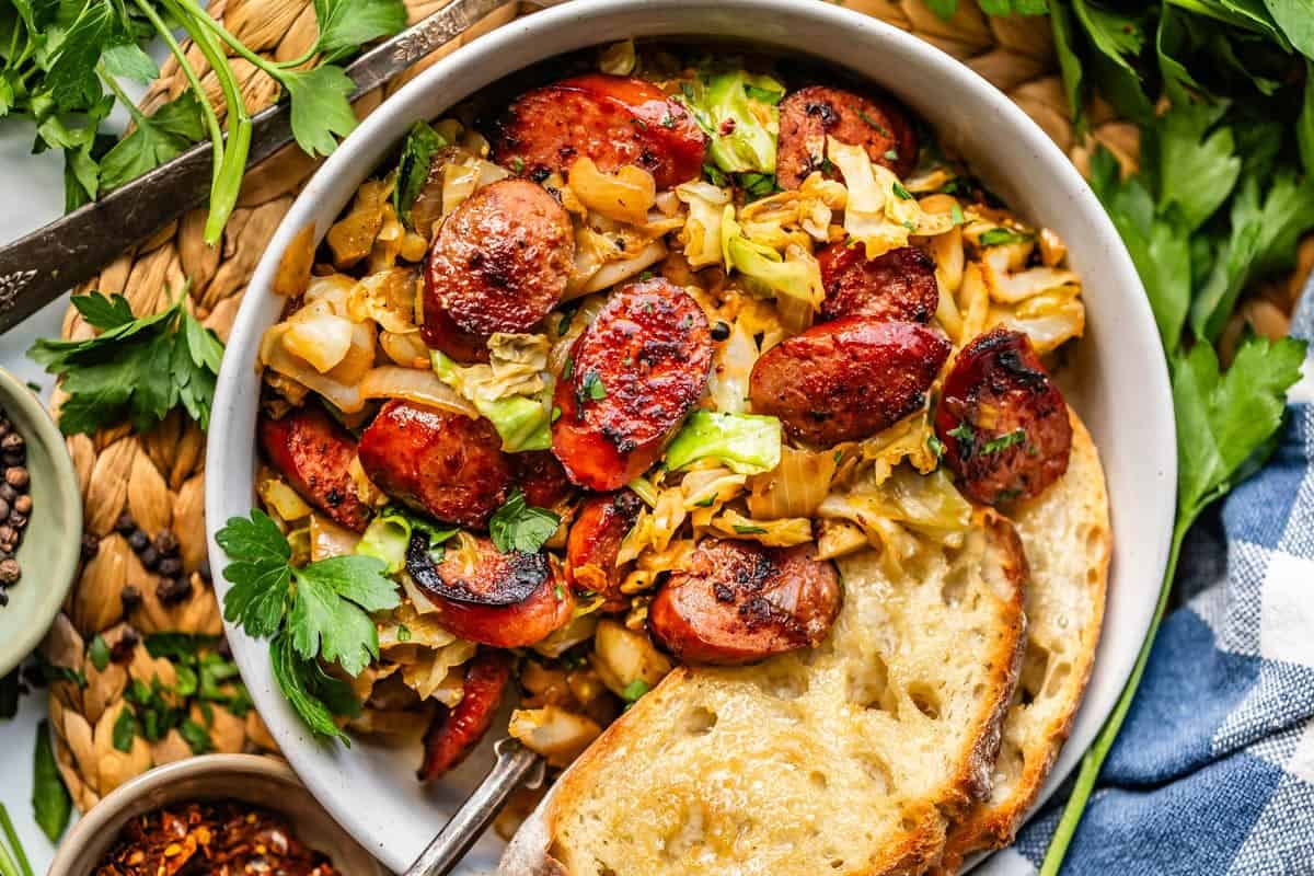 A bowl of saut&eacute;ed cabbage and sliced sausage garnished with parsley, served with two slices of toasted bread. Fresh herbs and seasonings are scattered around on a woven placemat.