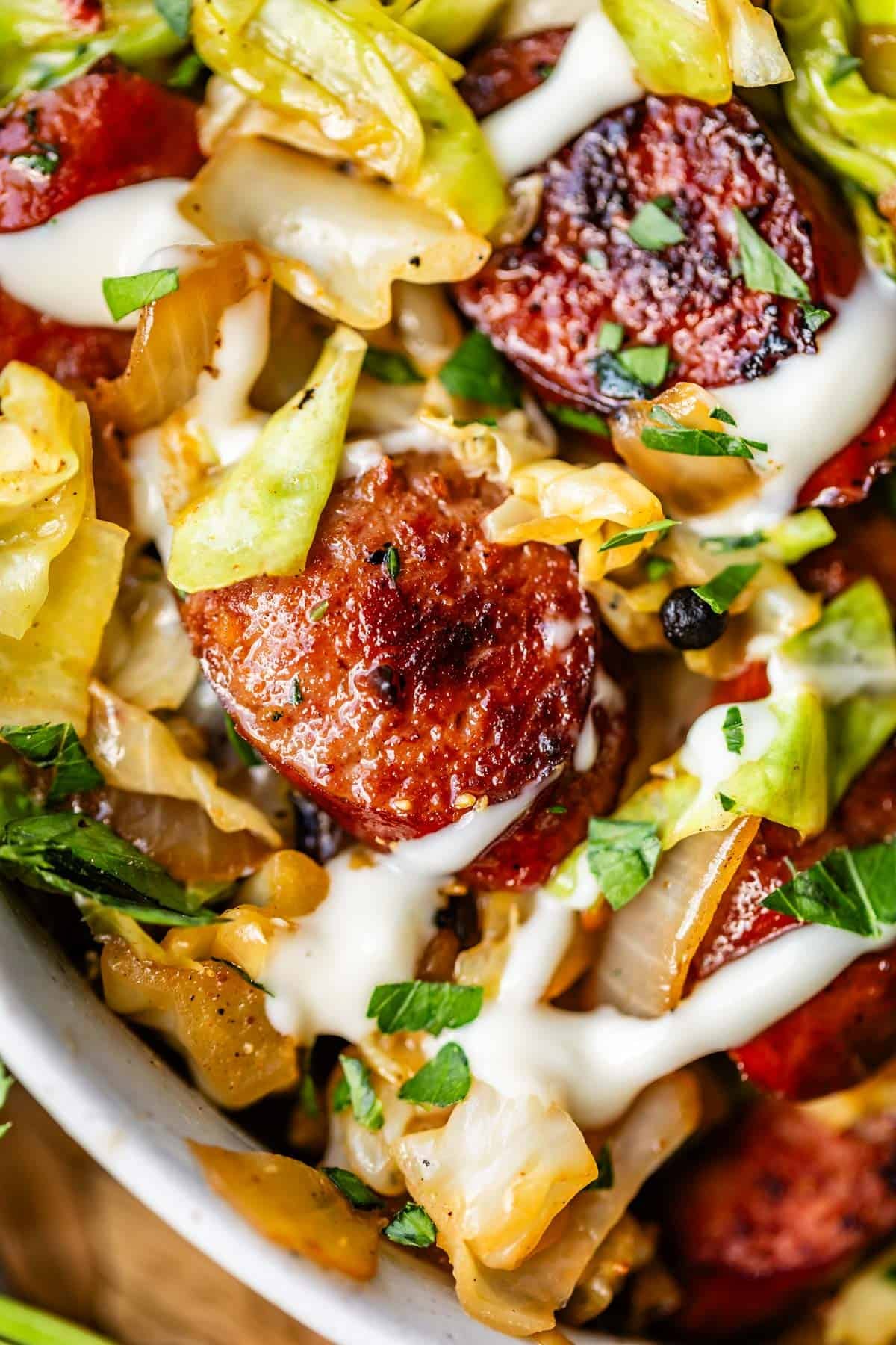 Close-up of saut&eacute;ed sausage slices with caramelized edges, cooked cabbage, and onions, topped with a drizzle of creamy sauce and sprinkled with chopped parsley.