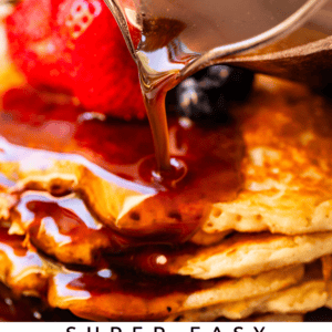 A close-up of maple syrup being poured from a jug onto a stack of pancakes topped with fresh strawberries and blueberries. Text reads: “Super Easy Homemade Maple Syrup – The Food Charlatan.”.