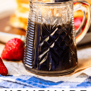 A glass pitcher of homemade maple syrup sits on a table next to fresh strawberries, with a stack of pancakes blurred in the background. Text reads: Homemade Maple Syrup. The Food Charlatan.