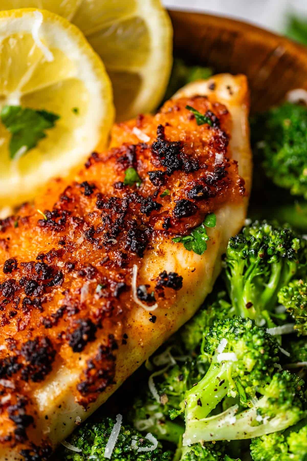 A close-up of a crispy, golden-brown chicken breast served with bright green broccoli florets and garnished with lemon slices and fresh herbs.