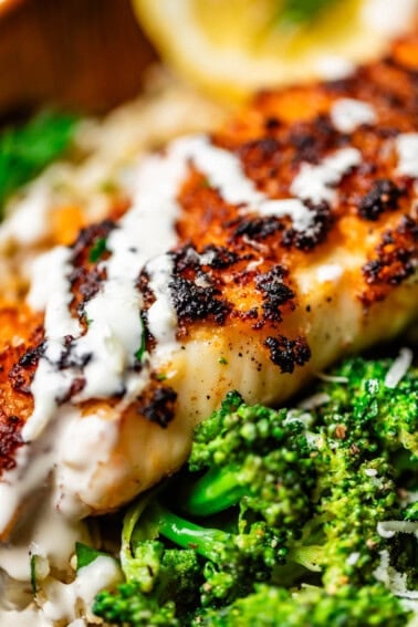 Close-up of a piece of browned, seasoned fish topped with a creamy white sauce, served alongside bright green broccoli and a slice of lemon in the background.