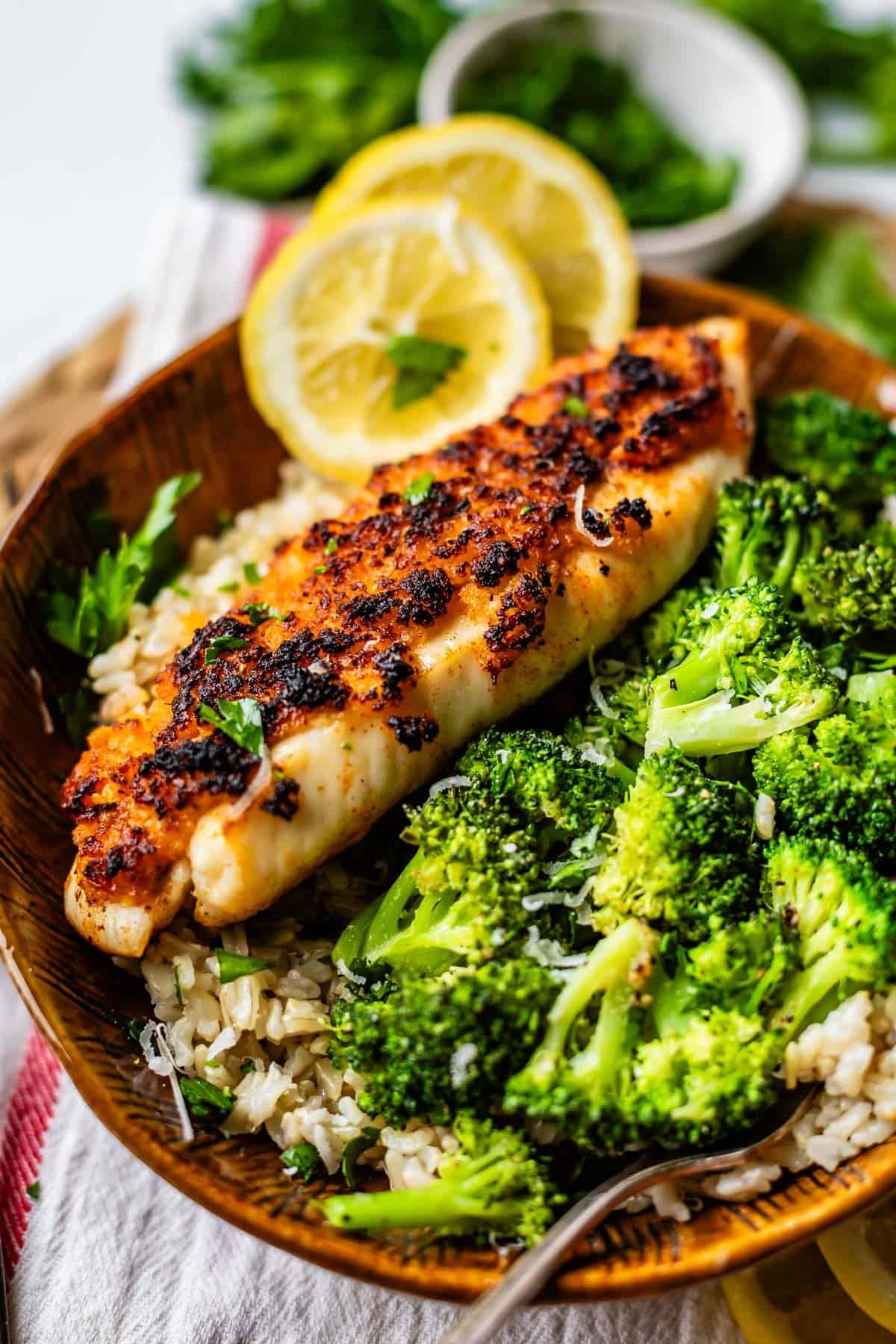 A bowl of rice topped with a grilled, seasoned fish fillet, steamed broccoli, and garnished with fresh parsley and lemon slices.