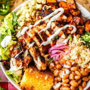 A colorful salad bowl topped with grilled chicken, corn, coleslaw, baked beans, pickled onions, lettuce, jalapeños, a golden cornbread slice, and drizzles of sauce, sitting on a checkered cloth.