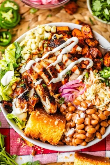 A colorful salad bowl topped with grilled chicken, corn, coleslaw, baked beans, pickled onions, lettuce, jalapeños, a golden cornbread slice, and drizzles of sauce, sitting on a checkered cloth.