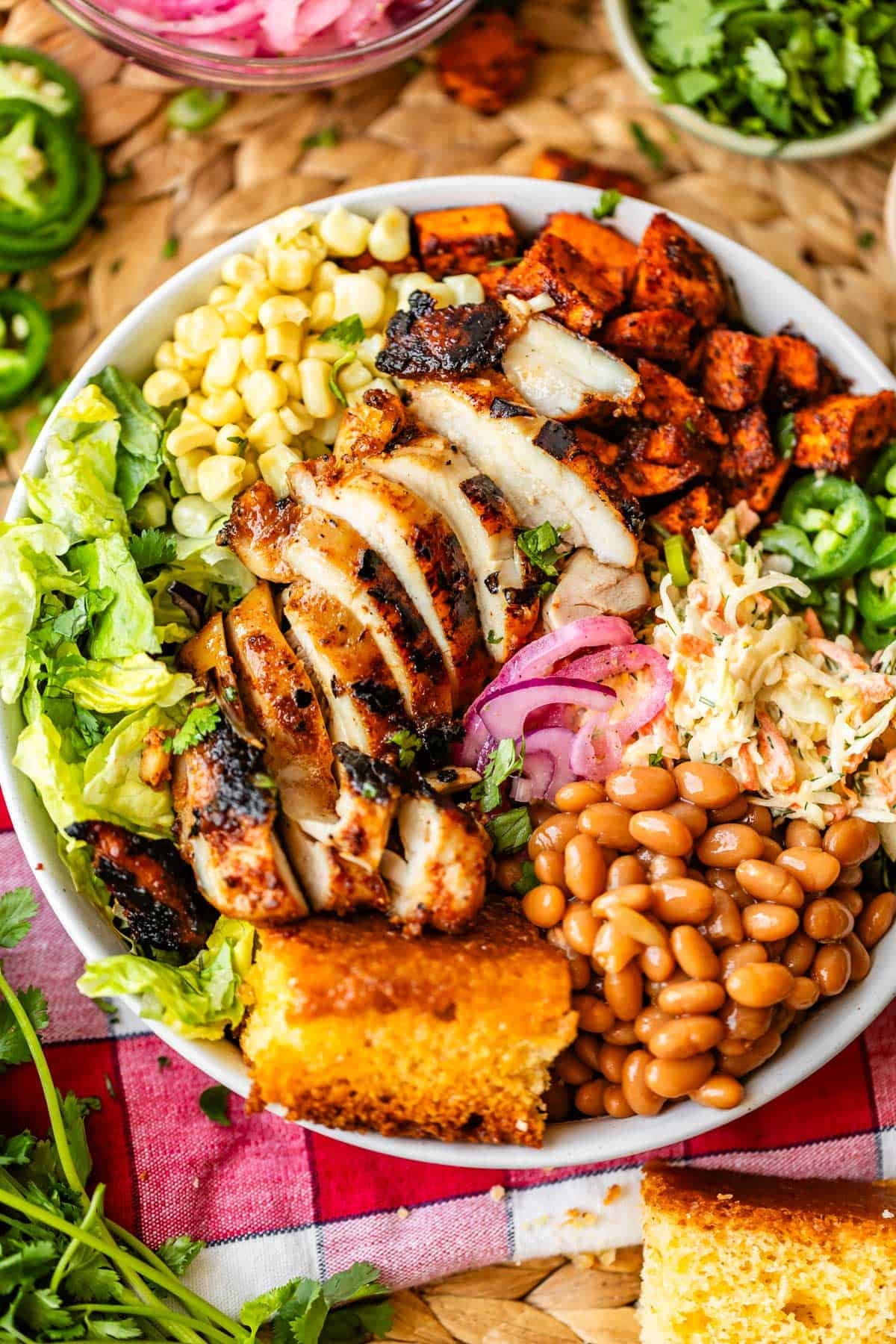 A bowl filled with sliced grilled chicken, corn, lettuce, roasted sweet potatoes, coleslaw, pickled onions, pinto beans, jalapeños, and a piece of cornbread, arranged on a colorful picnic cloth.