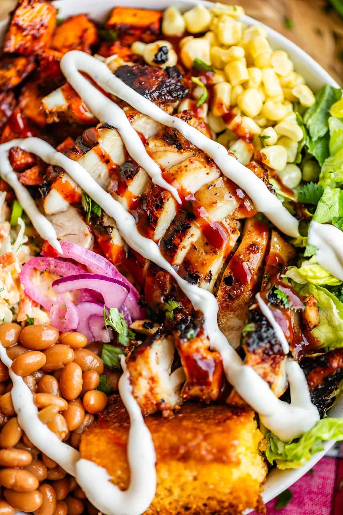 A colorful plate with grilled chicken drizzled with white sauce, surrounded by baked beans, cornbread, corn, lettuce, pickled onions, coleslaw, and roasted sweet potatoes.