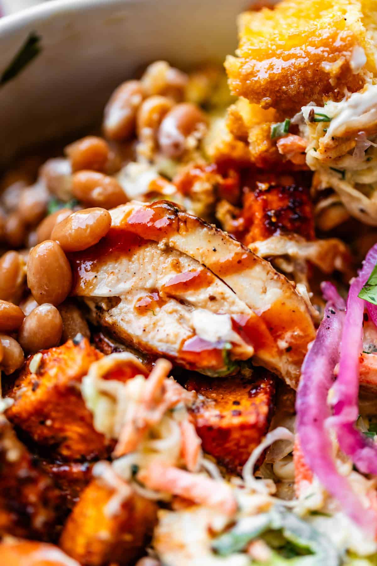 Close-up of a vibrant dish featuring grilled chicken slices, barbecue sauce, baked beans, cornbread, coleslaw, pickled red onions, and roasted sweet potatoes, all mixed together for a colorful and hearty meal.