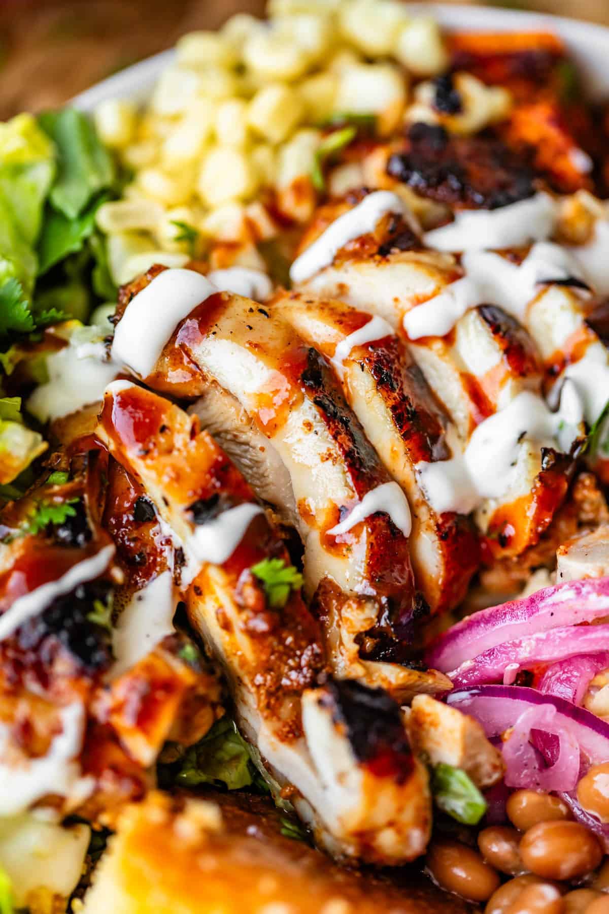 Close-up of a salad bowl with sliced grilled chicken drizzled with barbecue sauce and ranch dressing, surrounded by corn, lettuce, beans, pickled onions, and a slice of cornbread.