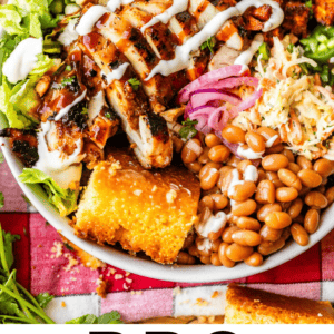 A bowl filled with BBQ chicken, beans, coleslaw, cornbread, red onions, lettuce, corn, rice, and a drizzle of creamy sauce, arranged on a colorful tablecloth. Text below reads BBQ Chicken Bowls - The Food Charlatan.