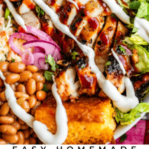 A close-up of a BBQ chicken bowl with grilled chicken, beans, corn, lettuce, pickled onions, and cornbread, all drizzled with ranch dressing. Text reads Easy Homemade BBQ Chicken Bowls - The Food Charlatan.