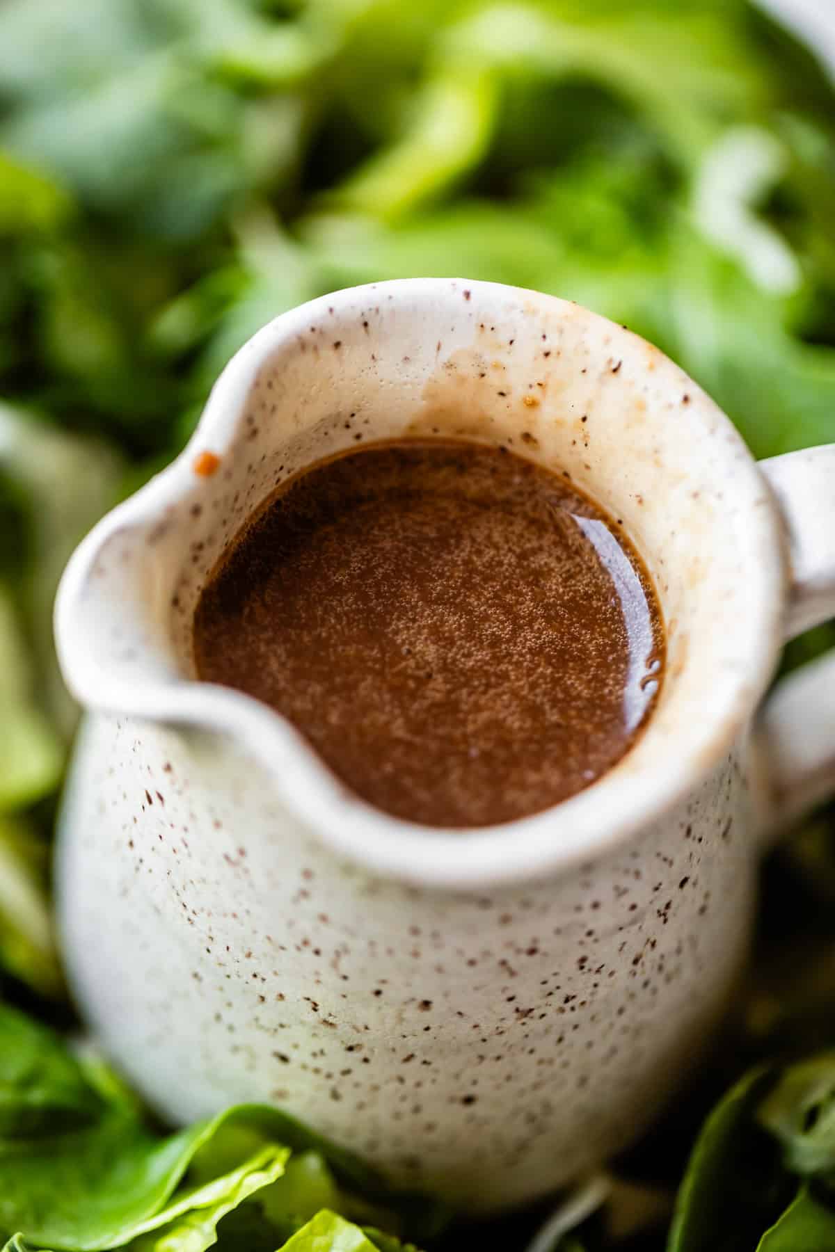 A close-up of a small ceramic pitcher filled with brown vinaigrette dressing, surrounded by fresh green leafy vegetables.