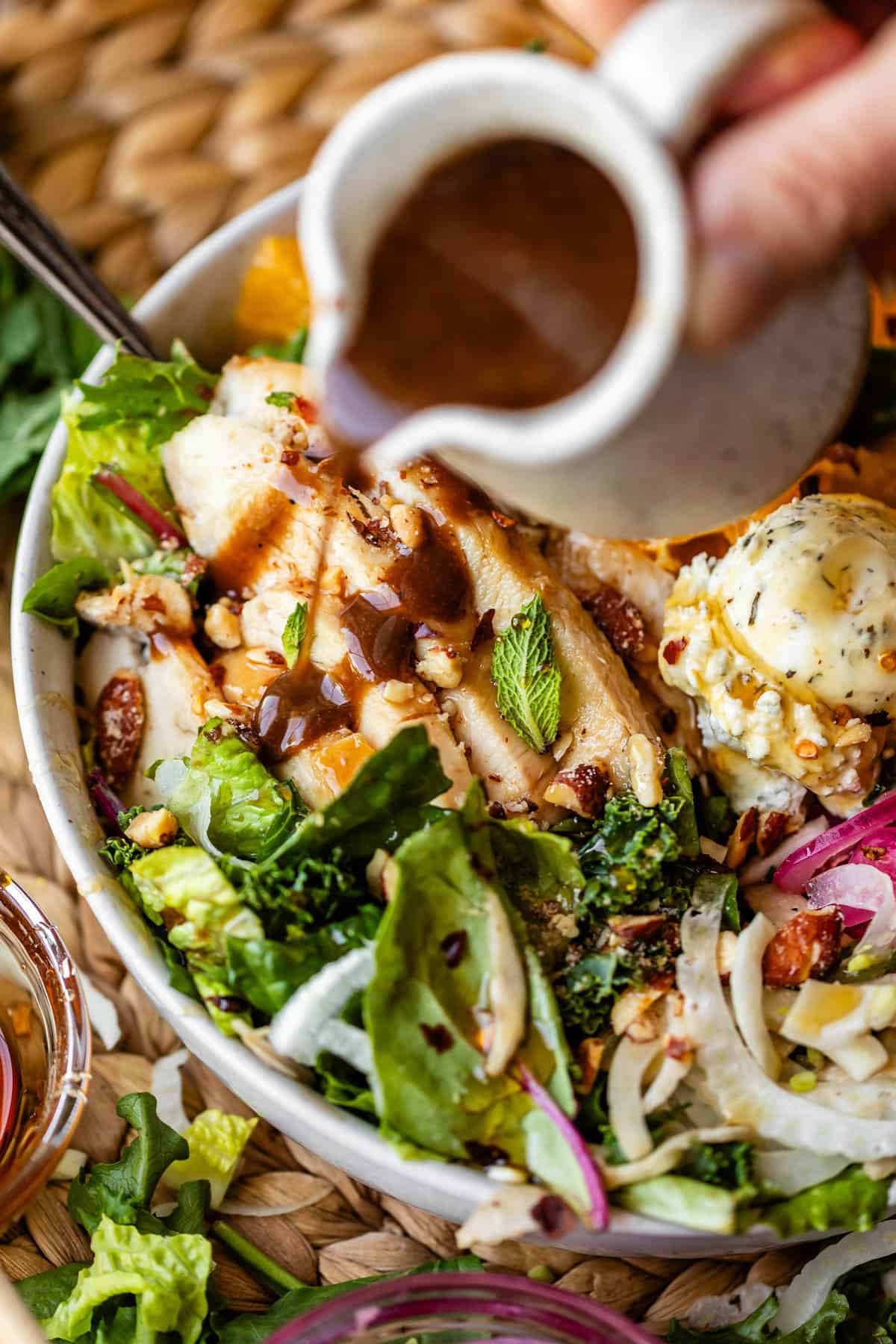 A close-up of a fresh salad with grilled chicken slices, leafy greens, red onions, and a cheese ball, as someone pours brown dressing from a small pitcher over the dish.