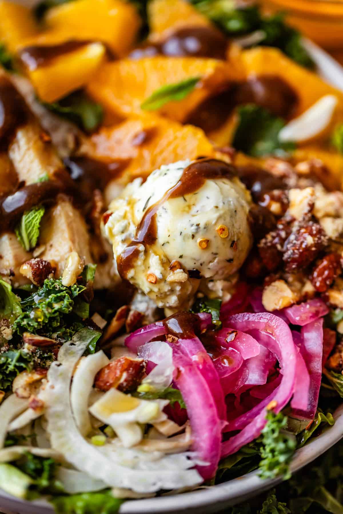 A close-up of a vibrant salad featuring leafy greens, orange slices, grilled chicken, pickled red onions, crumbled nuts, and a scoop of herbed cheese, all drizzled with a dark dressing.