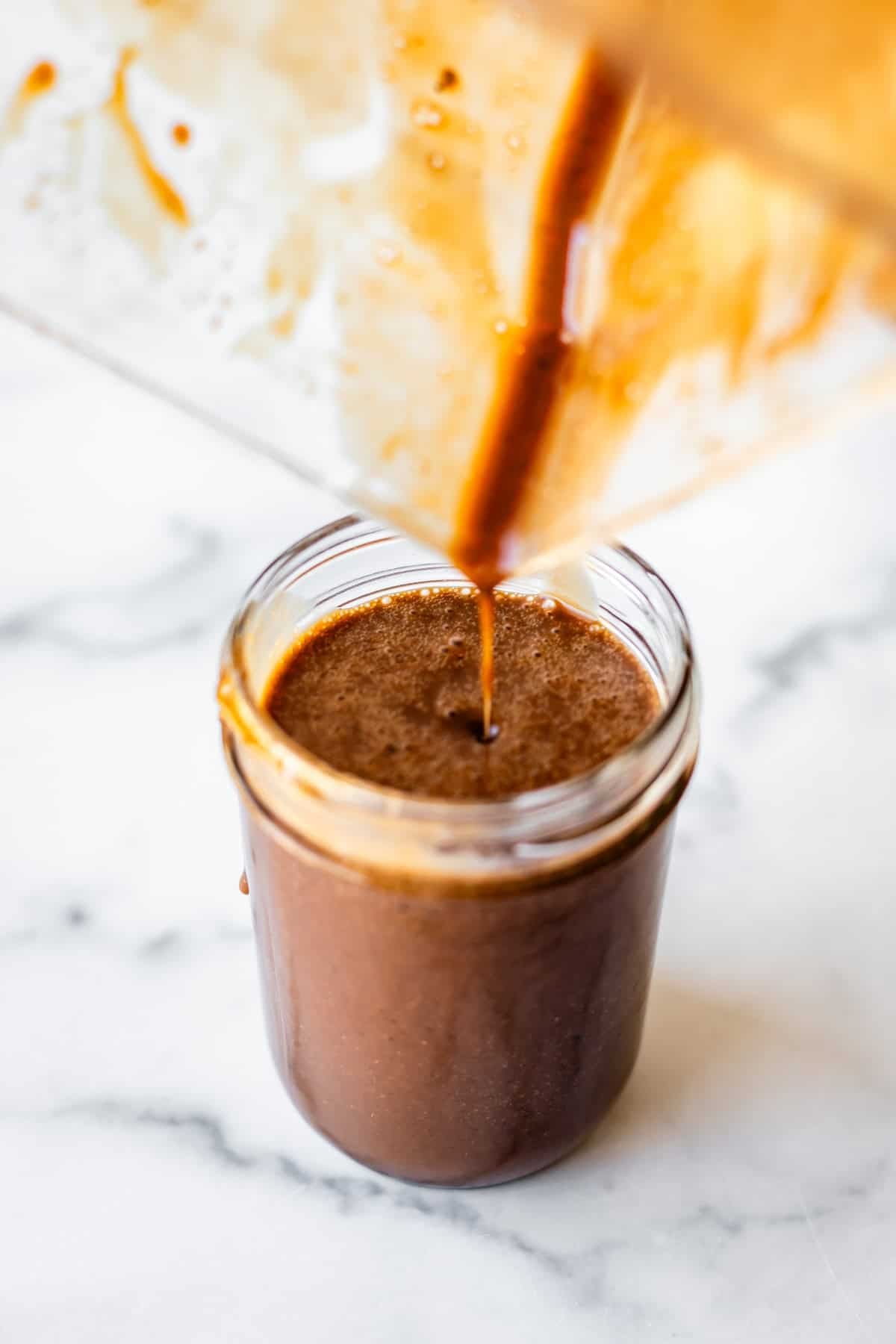 A thick, dark brown liquid is being poured from a blender into a glass jar on a white marble surface. Some of the liquid is visible inside the blender, and the jar is nearly full.
