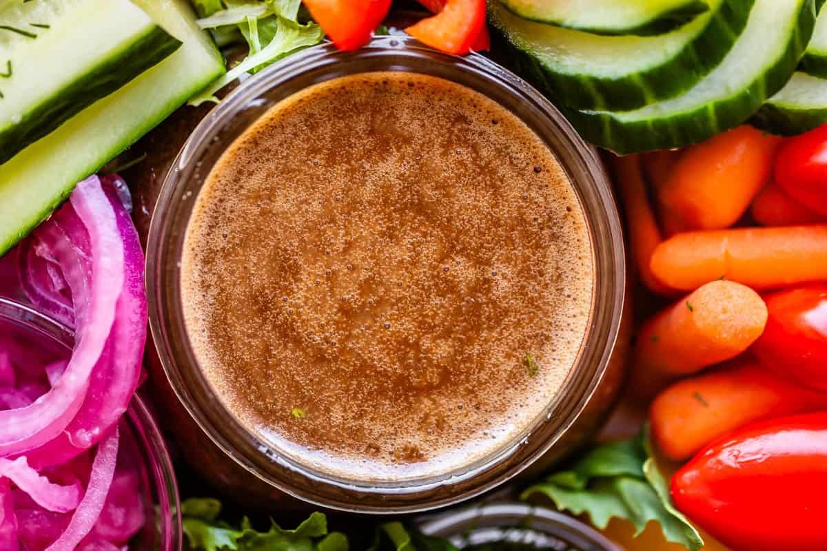 A close-up of a bowl of brown salad dressing surrounded by fresh vegetables, including sliced cucumbers, red onions, baby carrots, grape tomatoes, and red bell peppers.