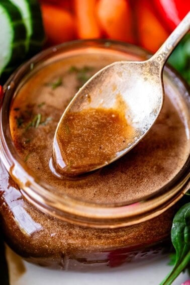 Close-up of a jar filled with brown vinaigrette dressing, with a spoon resting on top. Fresh vegetables, including cucumber and leafy greens, are visible around the jar.