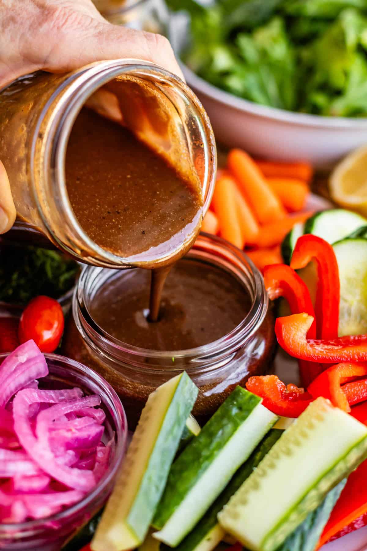 A hand pours dark brown salad dressing from a jar into a small bowl, surrounded by fresh vegetables including cucumbers, red bell peppers, carrots, lettuce, and pickled onions.