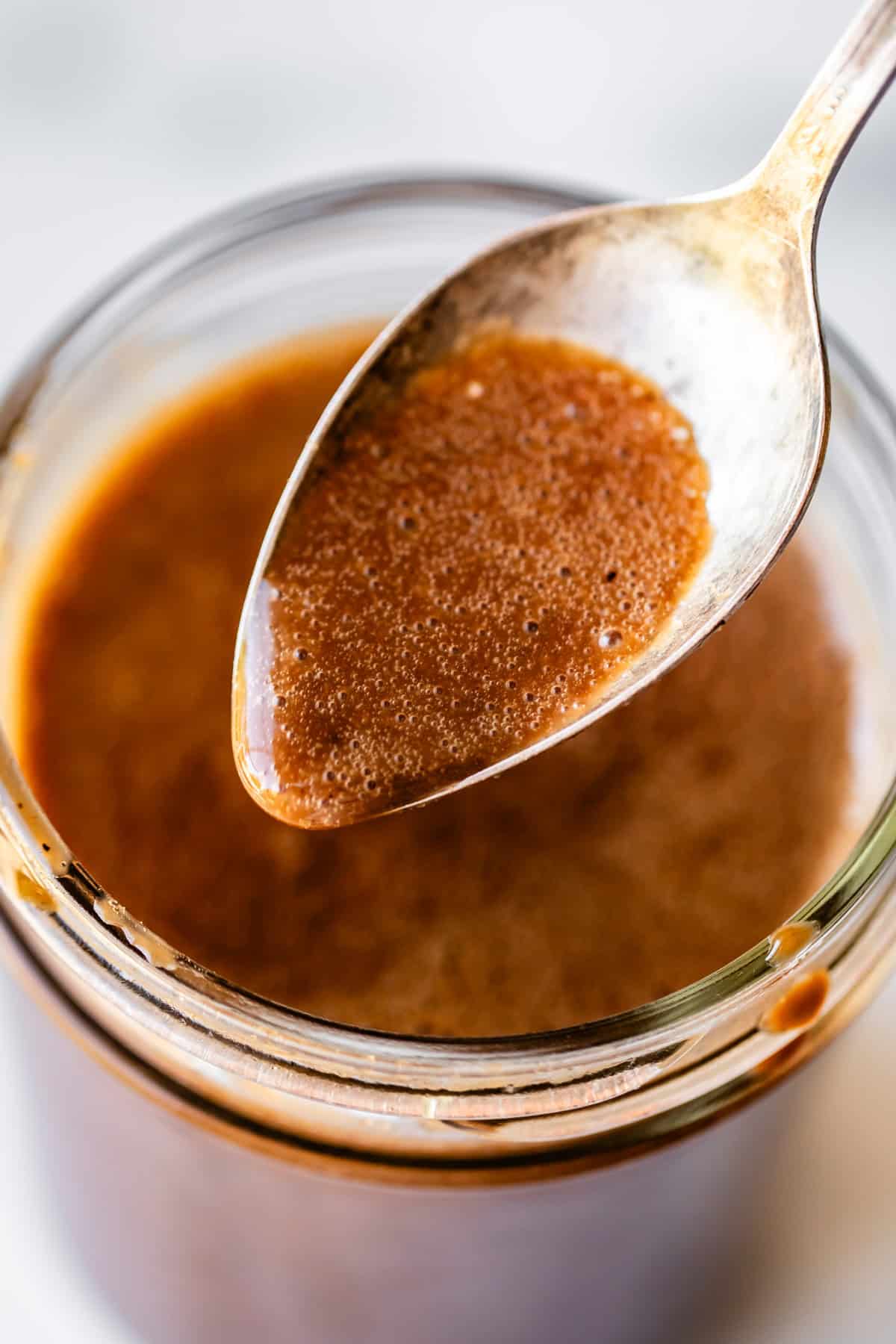 A close-up of a spoon dipped into a jar of thick, brown sauce or dressing, with the spoon held above the jar showing its smooth, glossy texture.
