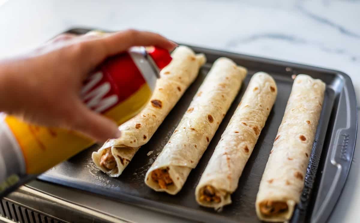A hand sprays nonstick cooking spray over four rolled tortillas filled with ingredients, placed on a baking sheet, ready to be baked.