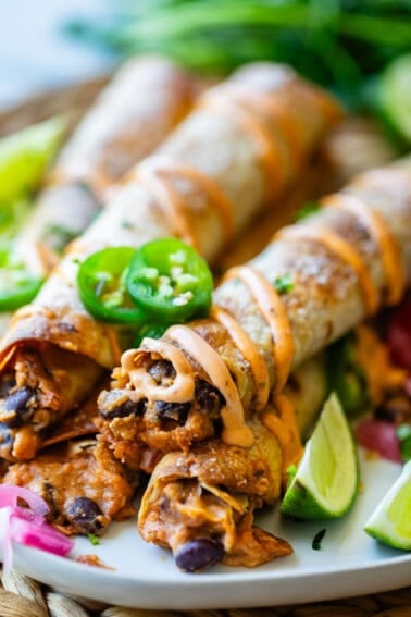 A close-up of crispy baked taquitos filled with black beans and sweet potatoes, topped with sliced jalapenos and a drizzle of orange sauce, served with lime wedges and pickled onions on a white plate.