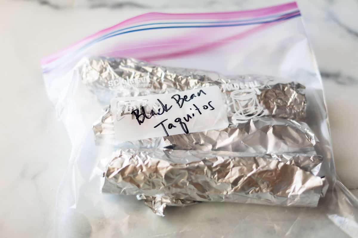 A clear plastic zip-top bag containing several foil-wrapped items labeled Black Bean Taquitos with handwritten text on white tape. The bag is placed on a light-colored surface.